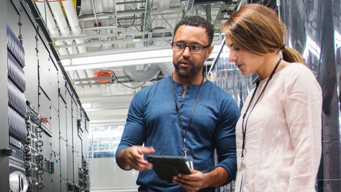 two IT professionals conversing in front of server racks, looking at a tablet screen